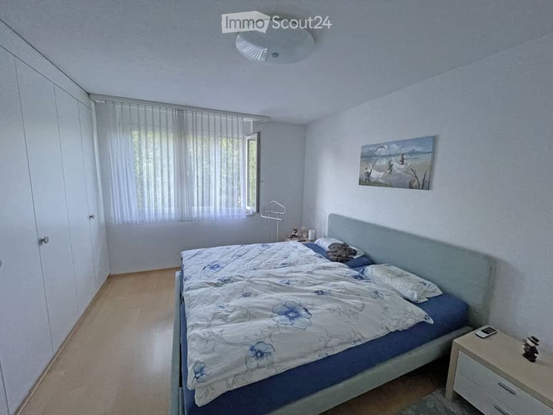 Bedroom with a double bed, nightstand, picture frame on the wall, and a wardrobe. The room has light-colored walls and a window with a curtain.