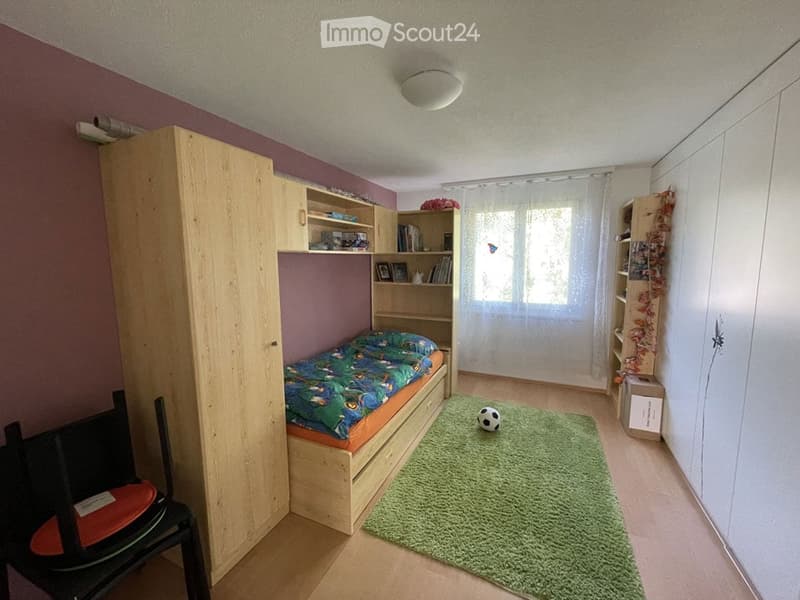 Cozy bedroom with a single bed, colorful bedding, wooden wardrobe, bookshelf, small green rug, and a black and white ball on the floor.