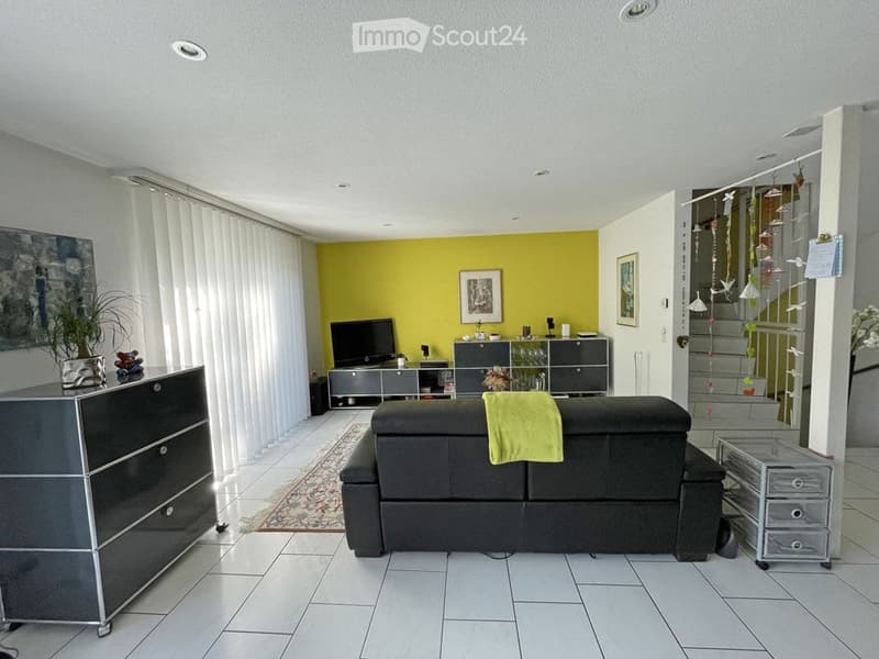 Modern living room with a black sofa, coffee table, TV stand, and a bright yellow accent wall. Features include a large window with white blinds, a small plant, and decorative picture frames.