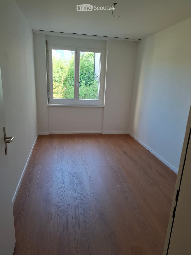 Empty room with wooden flooring, white walls, and a large window with white frames. The room has a door on the left side and a visible light fixture on the ceiling.