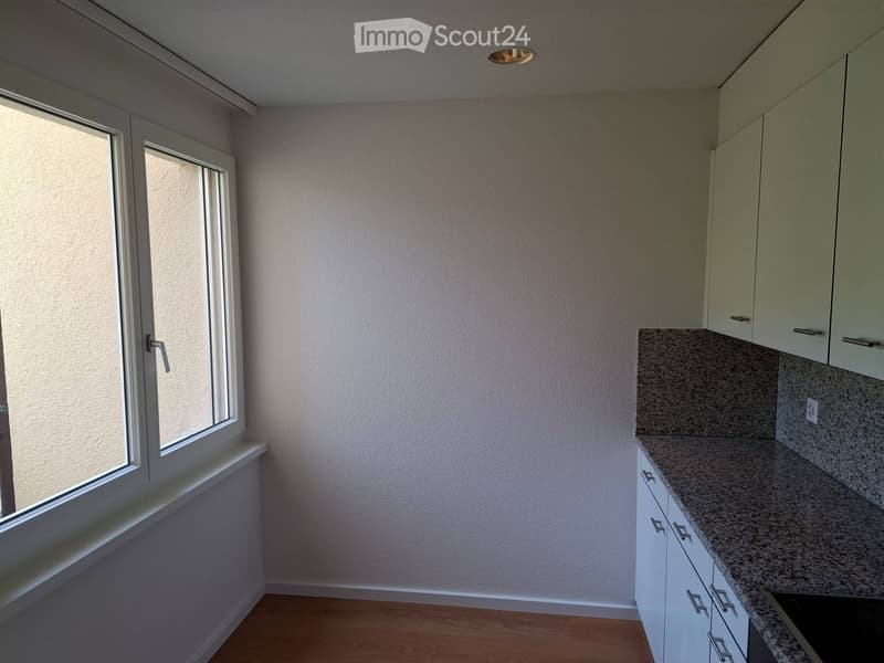Empty kitchen with light-colored cabinets, granite countertop, and wooden flooring. Large window allows natural light to enter.