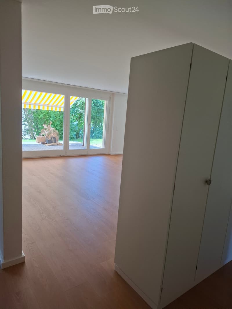 Empty room with wooden flooring, large sliding glass doors leading to a patio, and a view of greenery outside.