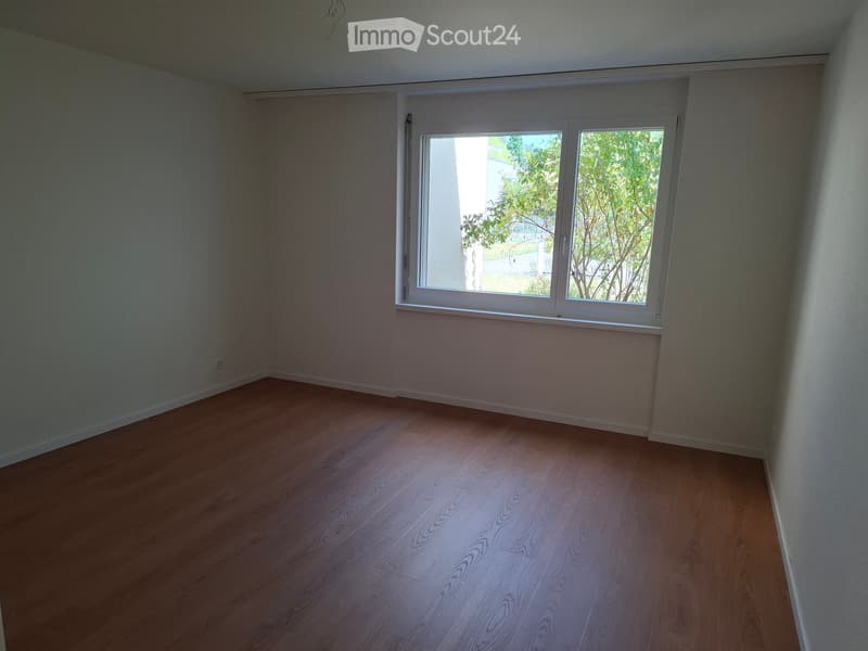 Empty room with wooden flooring and a large window letting in natural light. The walls are painted a neutral light color, and the room has no furniture or decorations.