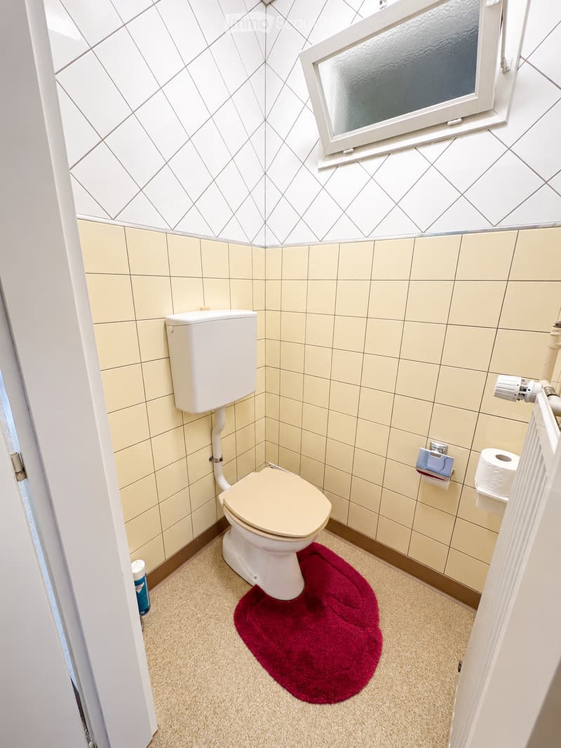 Small bathroom with beige tiled walls, a toilet, a toilet paper holder, and a red bath mat on the floor.