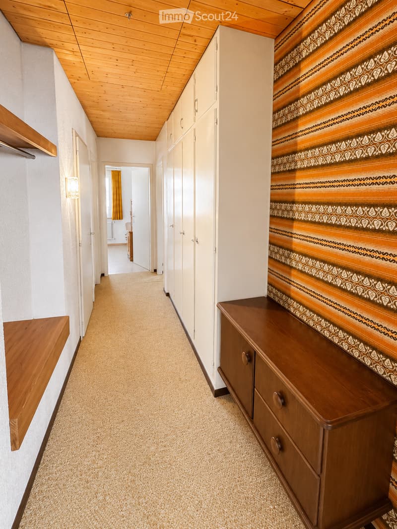 A narrow hallway with light wooden ceiling, white walls, carpeted floor, built-in white cabinets, and a wooden bench with drawers on the right side.