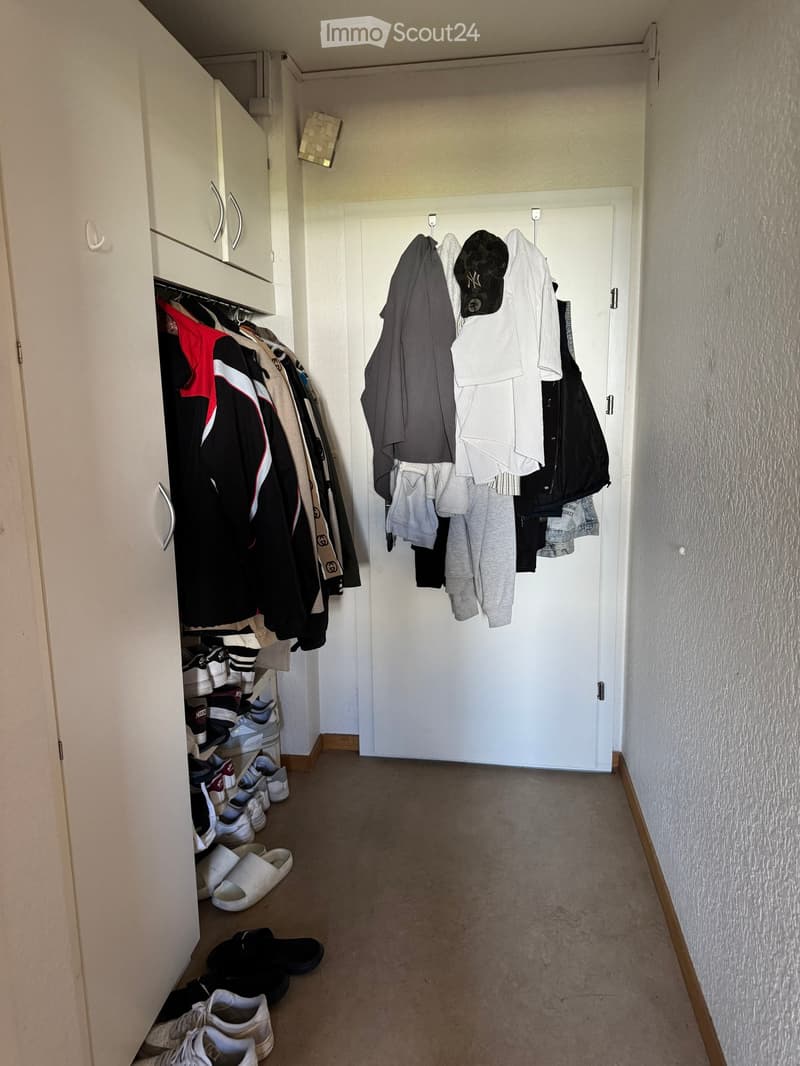 A narrow hallway with a carpeted floor, white walls, and a closed door at the end. Clothes are hanging on a rack, and shoes are placed on the floor near an open closet.