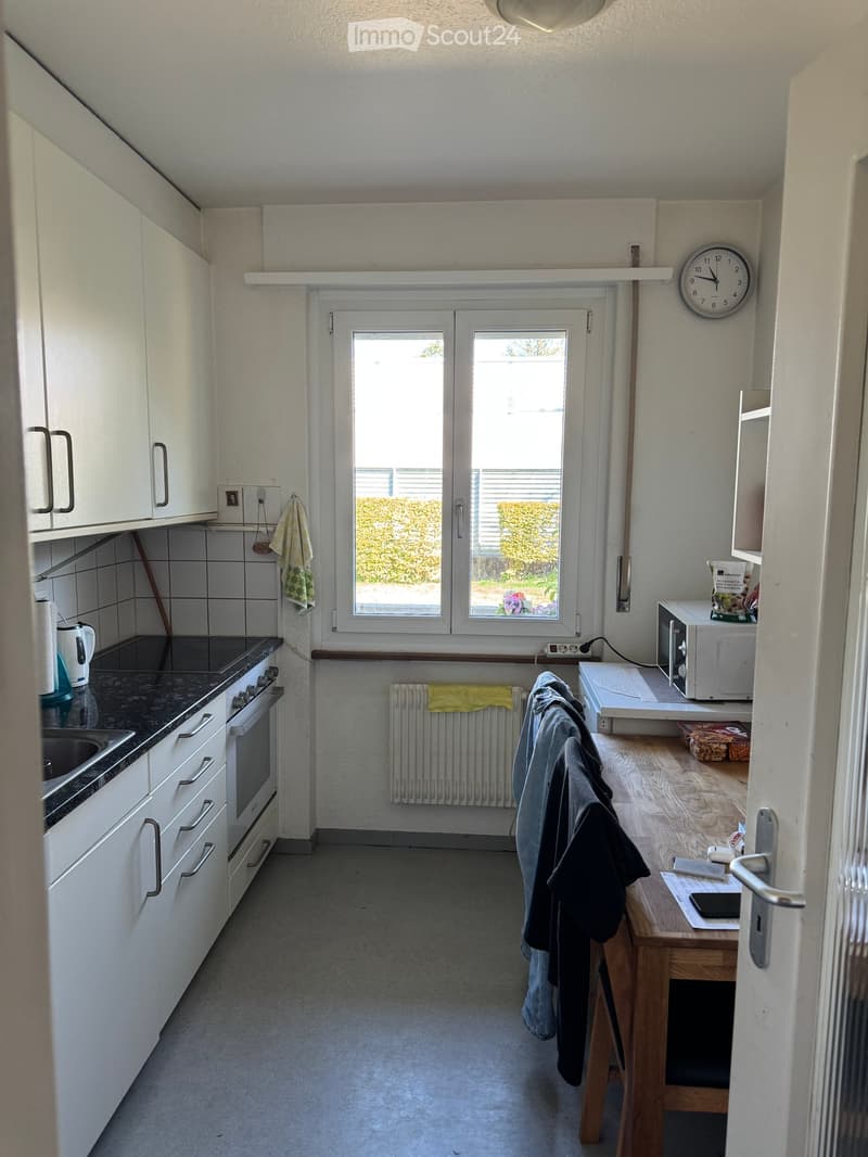 Modern kitchen with white cabinets, a black countertop, an oven, a sink, a microwave, a small table, and a clock on the wall. The room has a window with a view of greenery outside.