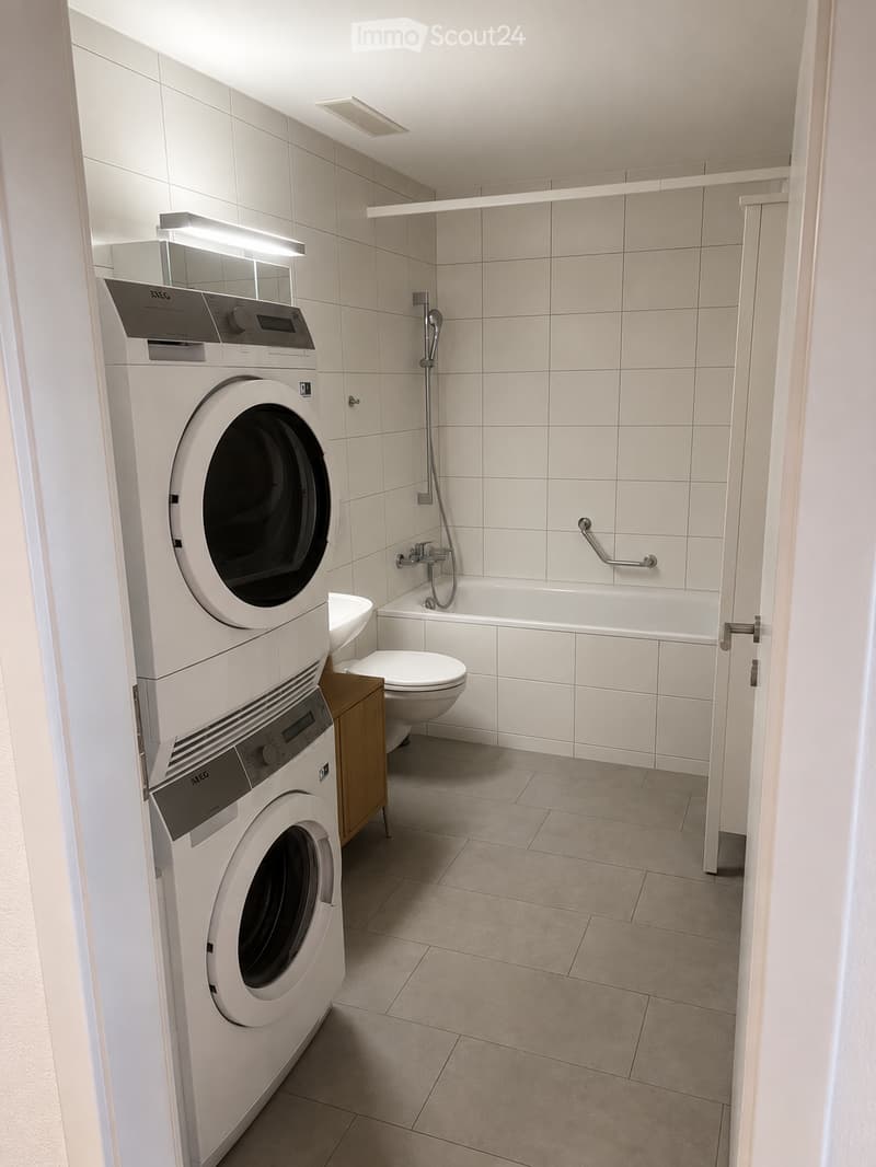 Modern bathroom with white tiles, a bathtub, toilet, sink, and stacked washing machine and dryer.