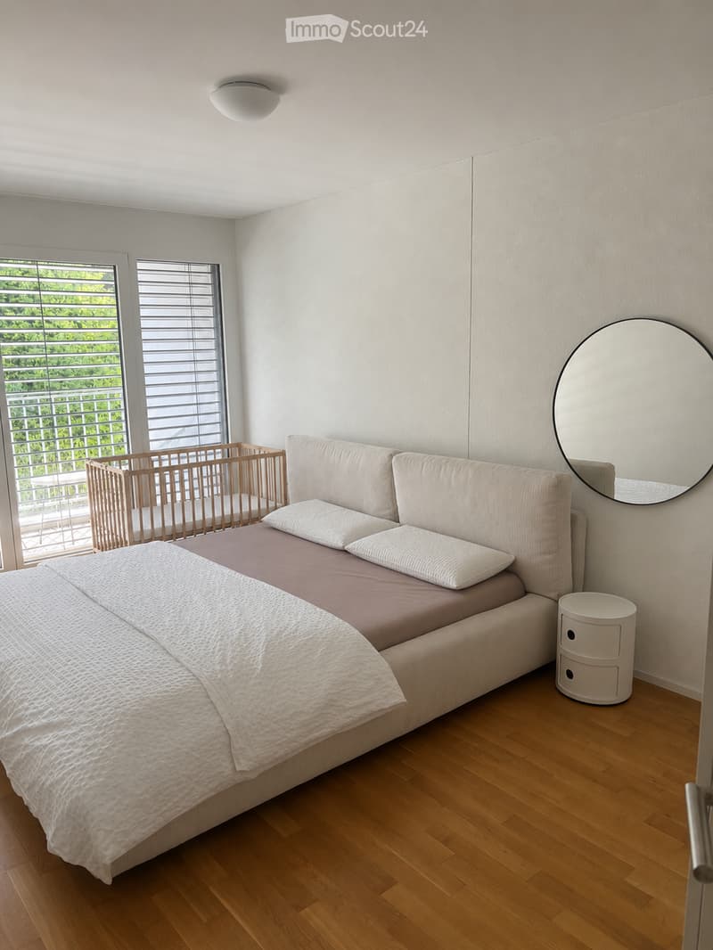 Modern bedroom with a large sofa bed, pillows, a round mirror, and a small nightstand. The room features wooden flooring and large windows with blinds, offering a view of greenery outside.