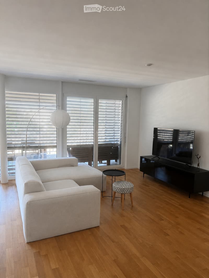Modern living room with a white sectional sofa, a black coffee table, a patterned stool, a floor lamp, a television on a black stand, and large windows with blinds.