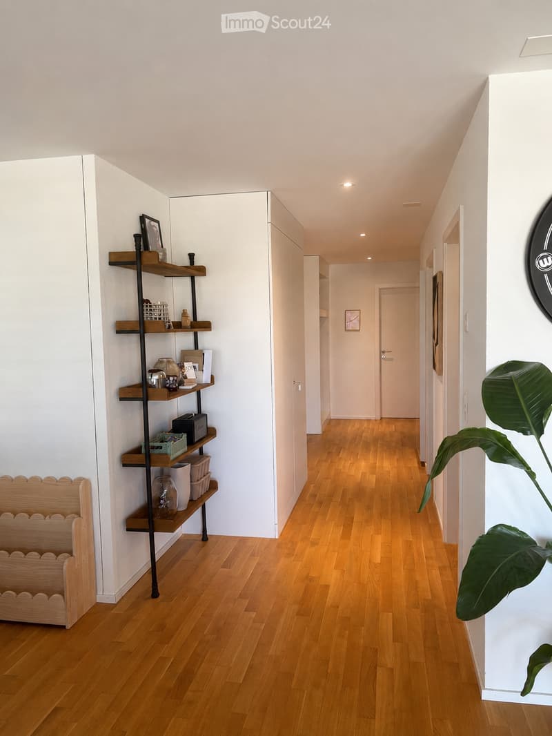 Modern hallway with light wooden flooring, white walls, and recessed ceiling lighting. Features a tall black shelving unit with decorative items and a wooden bench on the left, and a large leafy plant on the right.