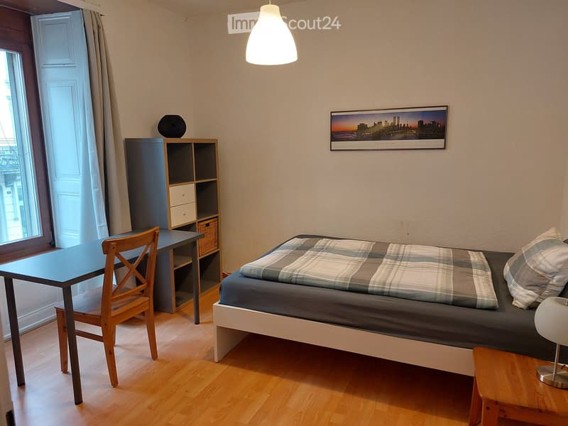 A modern, furnished bedroom with a single bed, a wooden chair, a small table, a shelf unit, and a hanging lamp.