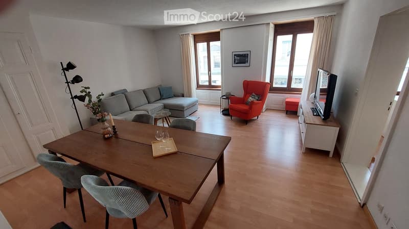 A modern living room with a grey sectional sofa, a red armchair, a wooden dining table with chairs, a television, and a floor lamp. The room features large windows with curtains and a view of the city.