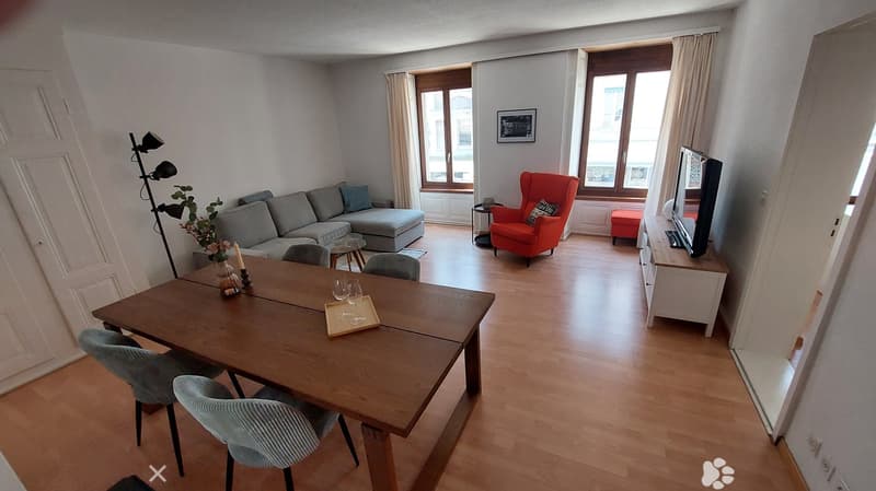 A modern living room with a grey sectional sofa, a red armchair, a wooden dining table with chairs, a television, and a floor lamp. The room features large windows with curtains and a view of the city.