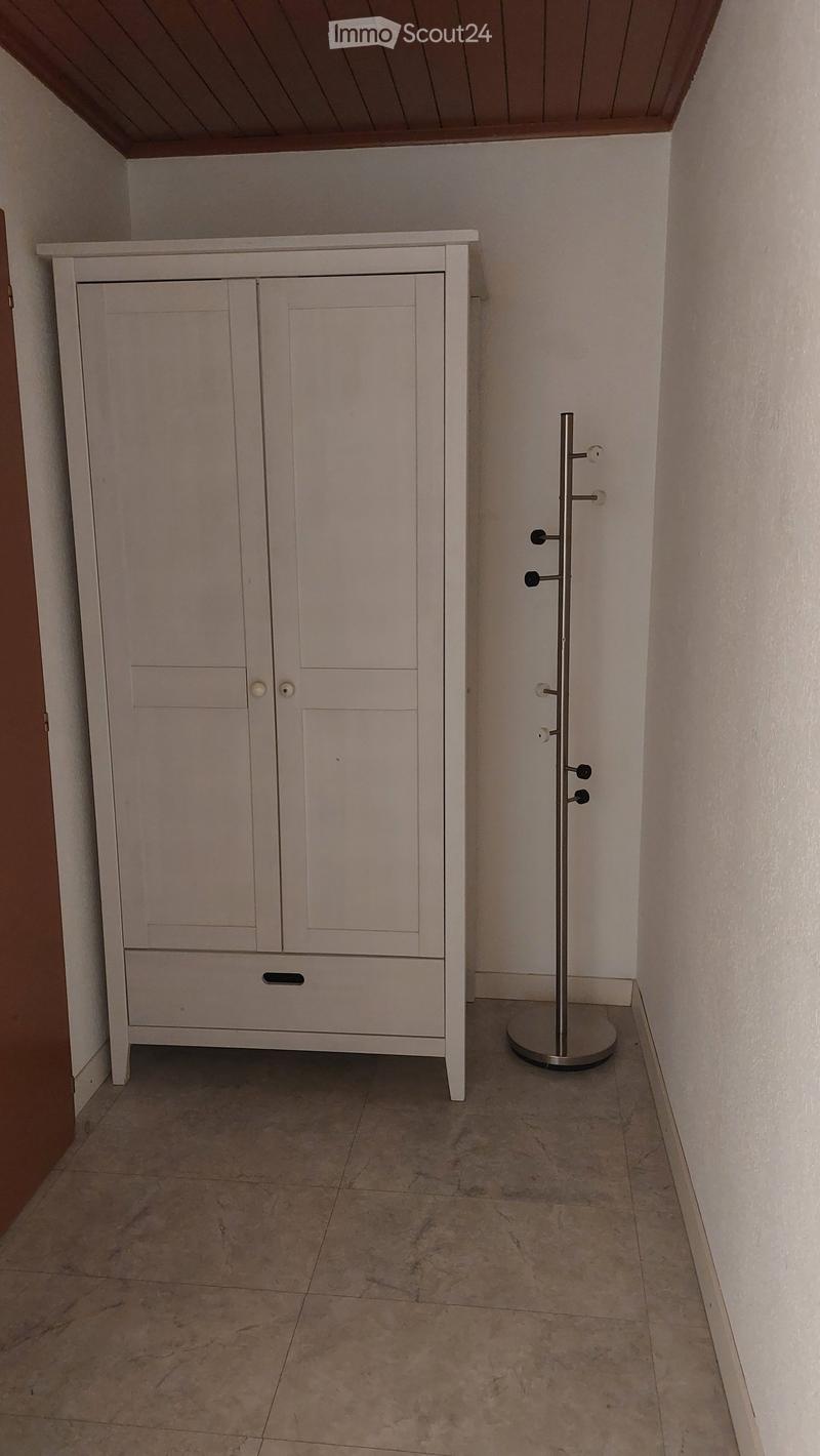 A narrow hallway with a white cabinet and a coat rack on the right side. The floor is tiled, and the walls are painted white.