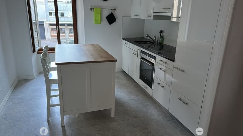 small kitchen, with a white island and grey tiled floor. white cabinets and a white counter. black countertops. kitchen appliances include sink, faucet, oven.