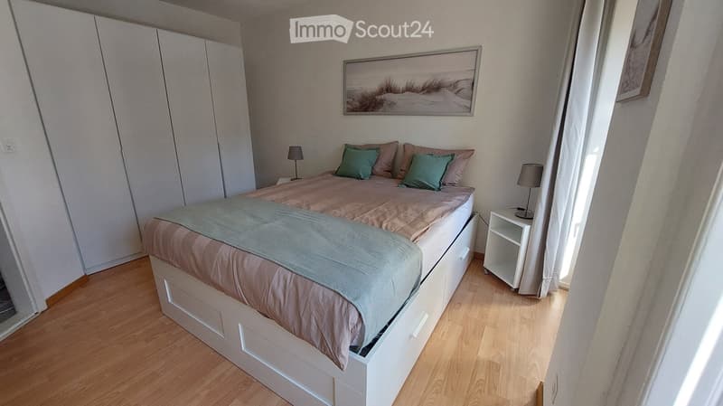Modern bedroom with a large bed, nightstands, lamps, and a wardrobe. The room features wooden flooring and large windows with white curtains.