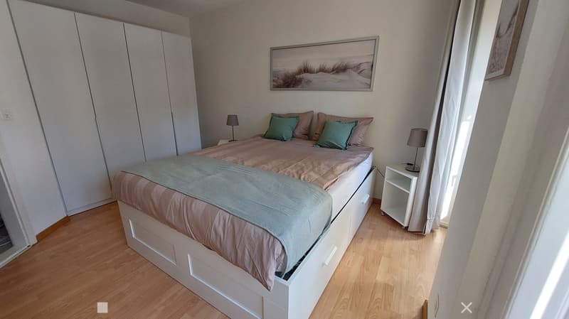 Modern bedroom with a large bed, nightstands, lamps, and a wardrobe. The room features wooden flooring and large windows with white curtains.