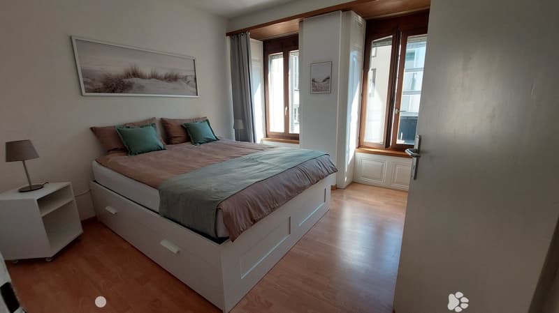 Bedroom with double bed, wooden floor, nightstand with lamp, white walls, two large windows, built-in wardrobe, picture frames on the wall.