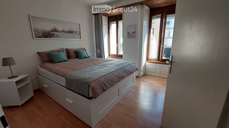 Bedroom with double bed, wooden floor, nightstand with lamp, white walls, two large windows, built-in wardrobe, picture frames on the wall.