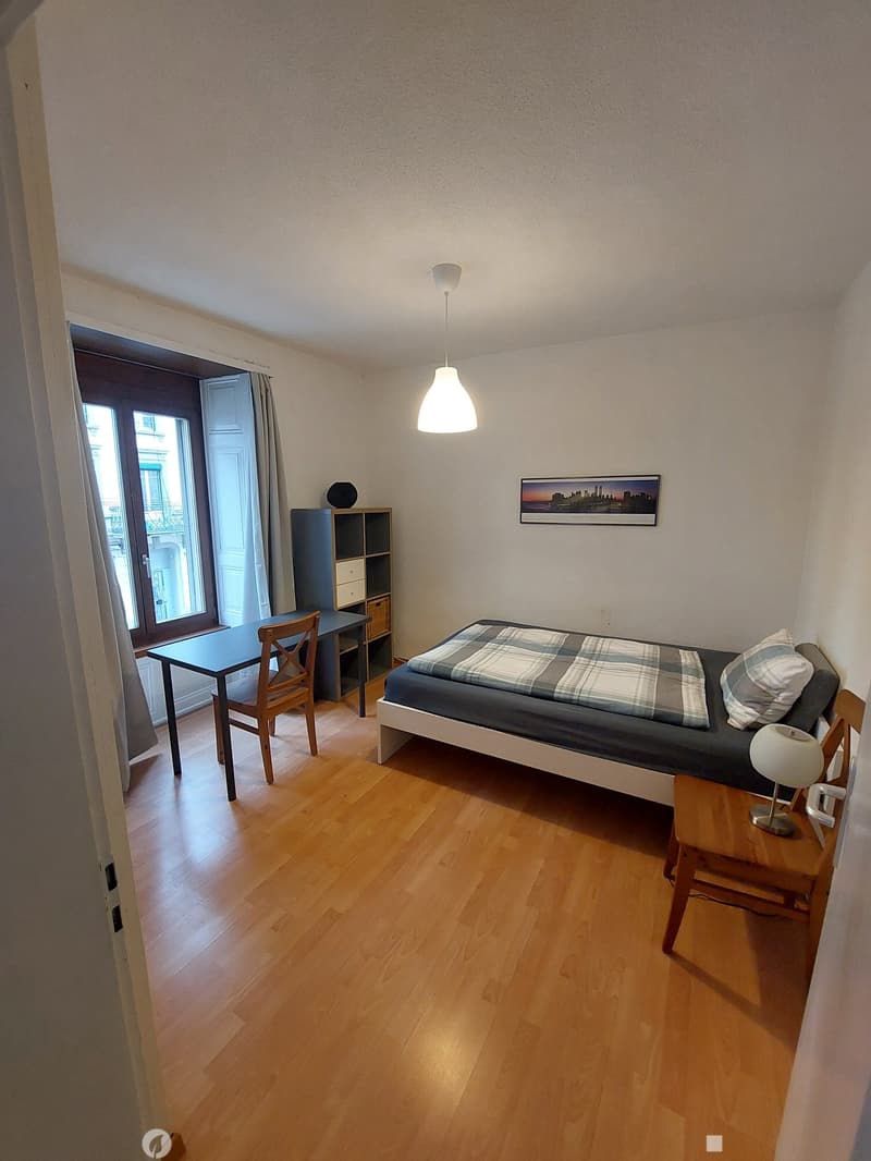 Cozy bedroom with wooden flooring, a single bed with plaid bedding, a small table with a lamp, a chair, and a cabinet with drawers.