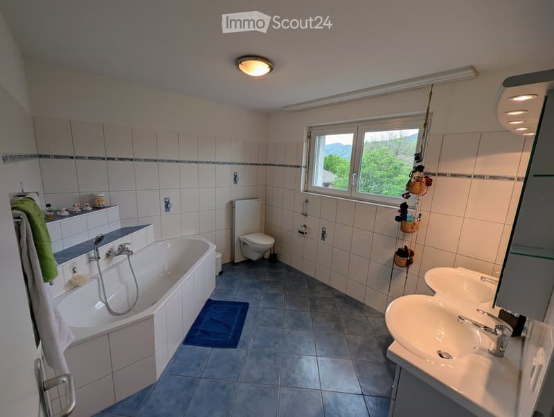Modern bathroom with a bathtub, double sink, toilet, and large window offering a scenic view. Features tiled walls, a towel rack, and a blue bath mat.