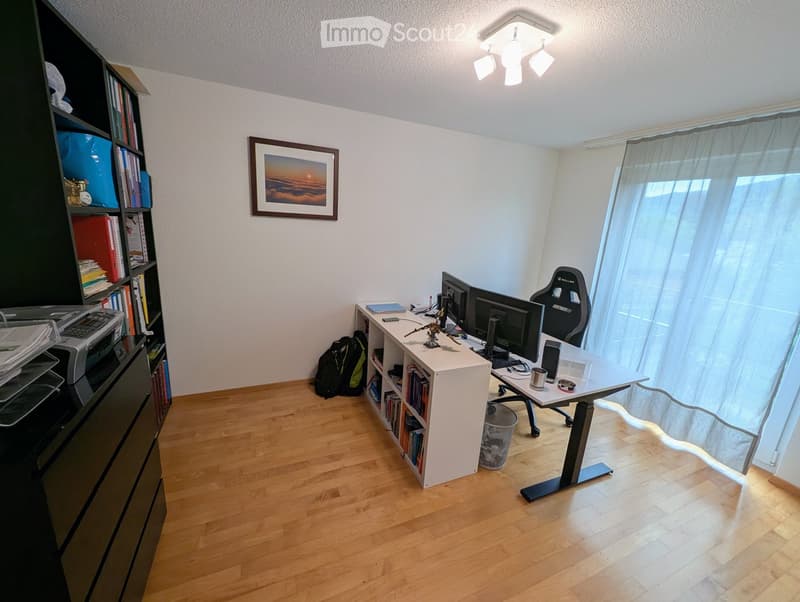 A well-organized home office with a wooden floor, large window with curtains, a standing desk with a chair, a bookshelf, and a printer.