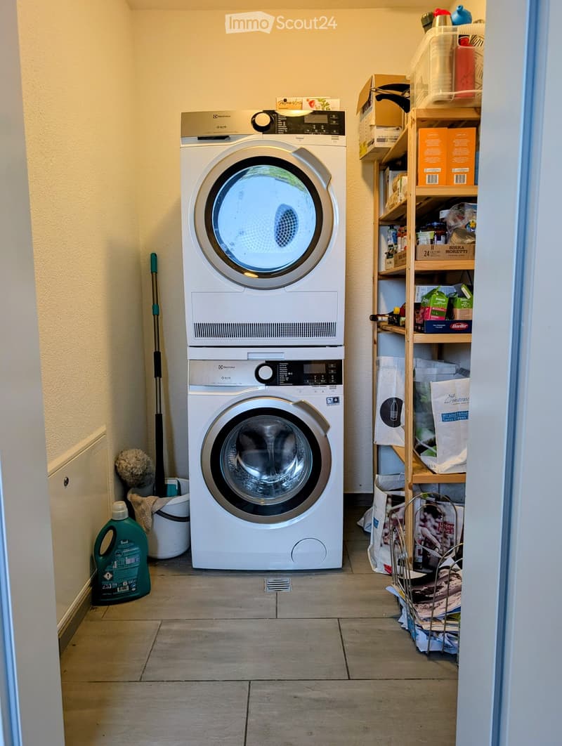 A small bathroom with a stacked washing machine and dryer, a wooden shelf with various items, a green bottle, and a mop leaning against the wall.