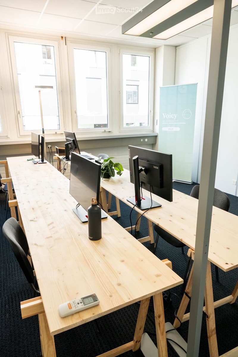 A modern office space with a long wooden table, multiple computer screens, chairs, a potted plant, and a water bottle on the table.