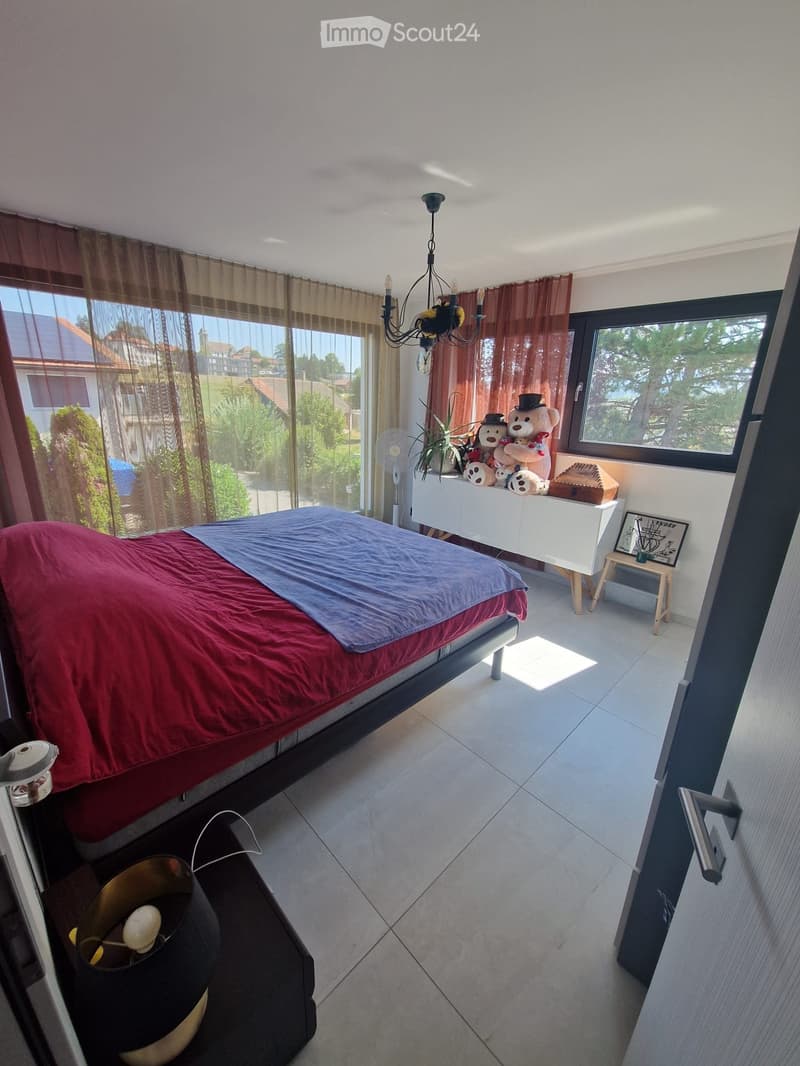 Bright bedroom with large windows, a bed with red and blue bedding, a chandelier, a small table with a fan, and decorative items on a white cabinet.