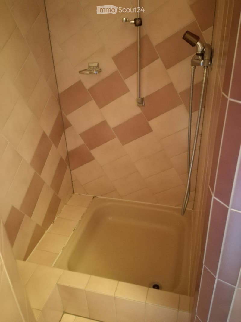 Shower area with beige tiled walls and floor, featuring a handheld showerhead and hose.