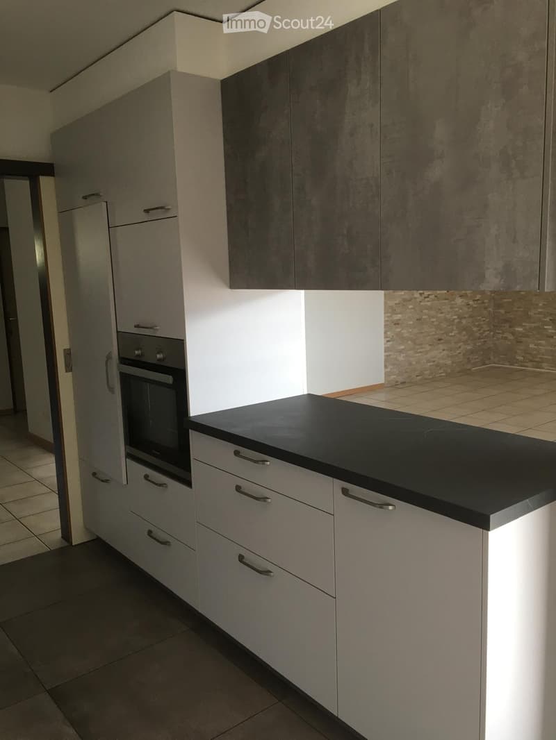 Modern kitchen with grey cabinets, black countertop, and built-in oven. Features tiled flooring and a small seating area visible in the background.