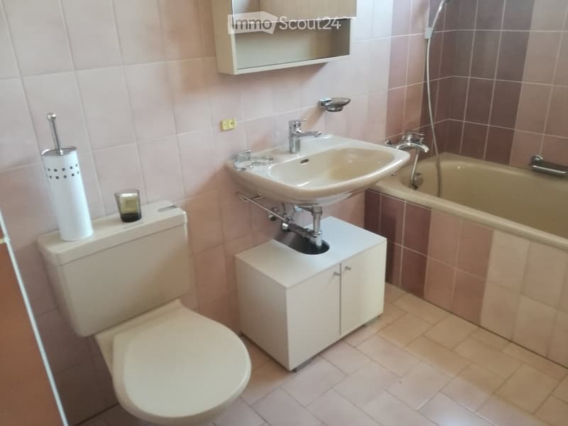 Bathroom with a toilet, sink, bathtub, and a small cabinet. The walls are tiled in light pink and brown, and the floor is tiled in beige.