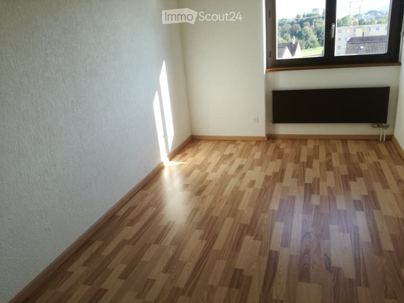Empty room with light wooden flooring, white walls, and a large window with a view of the outside. A radiator is mounted on the wall near the window.