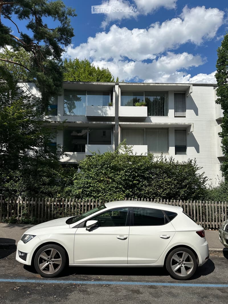 Modern two-story building with white facade, large windows, and balconies, surrounded by greenery and a parked car.