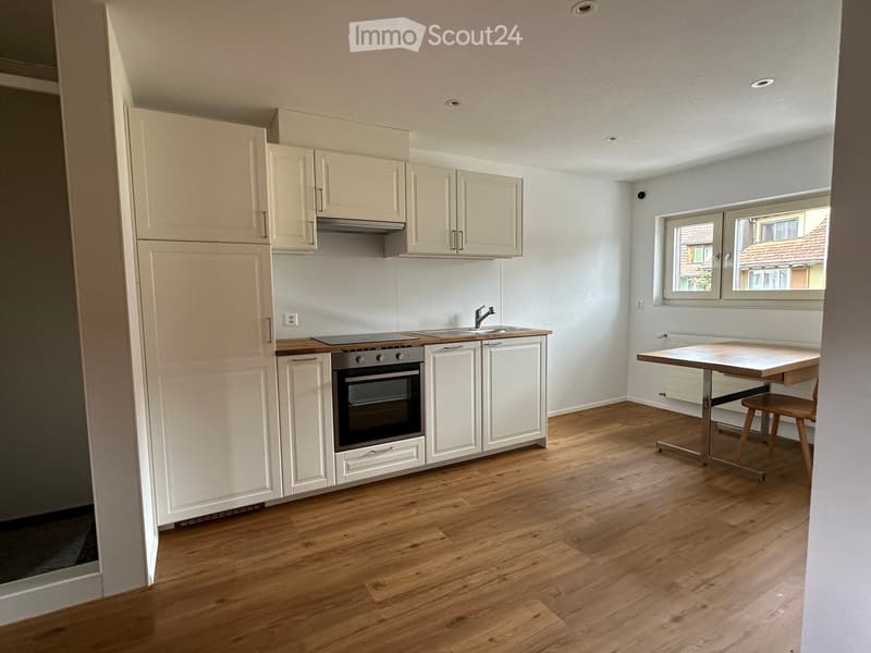 Modern kitchen with light-colored cabinets, wooden flooring, and a small dining area with a table and chair.