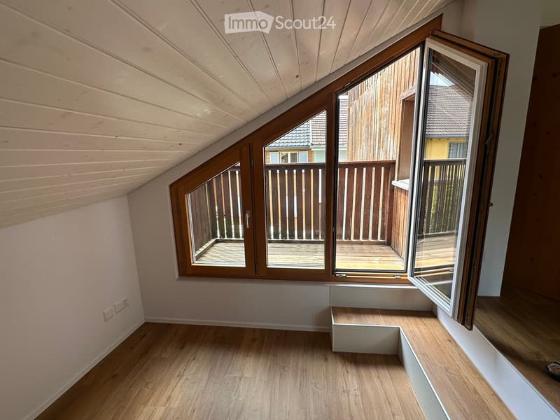 An empty attic room with wooden floors, white walls, and large windows opening to a wooden balcony. The ceiling has exposed wooden beams.