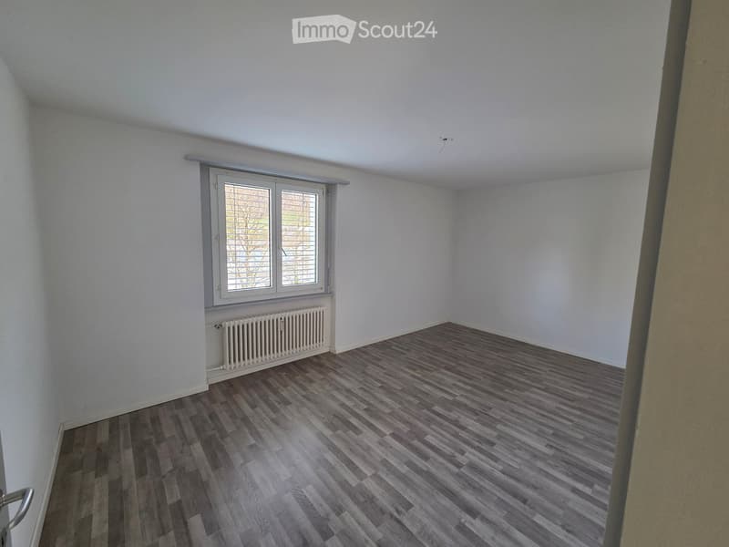 Empty room with wooden flooring, white walls, and a window with blinds. A radiator is visible below the window.