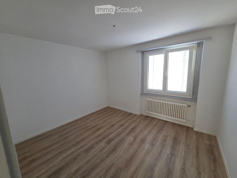 Empty room with light-colored walls, wooden flooring, and a window with blinds. A radiator is visible below the window.