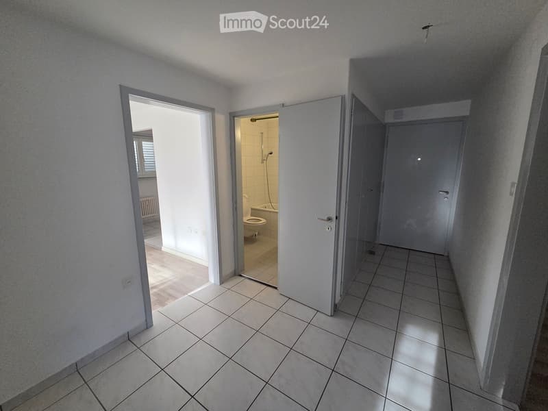 A clean, tiled hallway with white walls, leading to a partially open door revealing a bathroom with a toilet, bathtub, and shower. The hallway has a simple design with a window on the left side.
