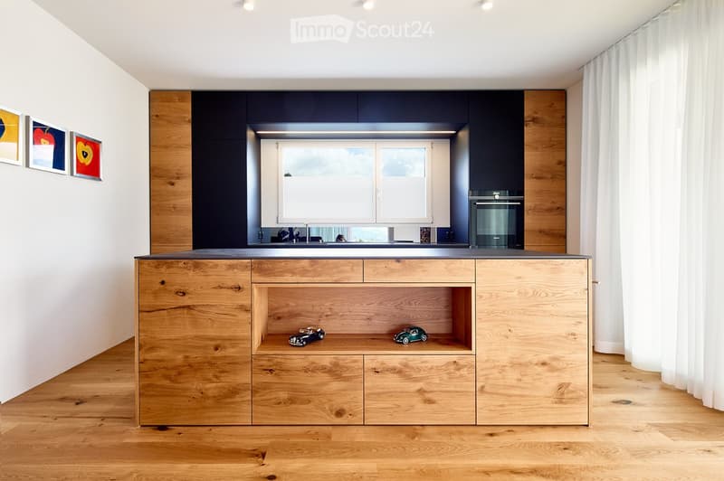 Modern kitchen with wooden cabinets, a central island, and a window overlooking a scenic view. The island features drawers and open shelving with decorative toy cars. The kitchen includes a sink, oven, and a sleek faucet.