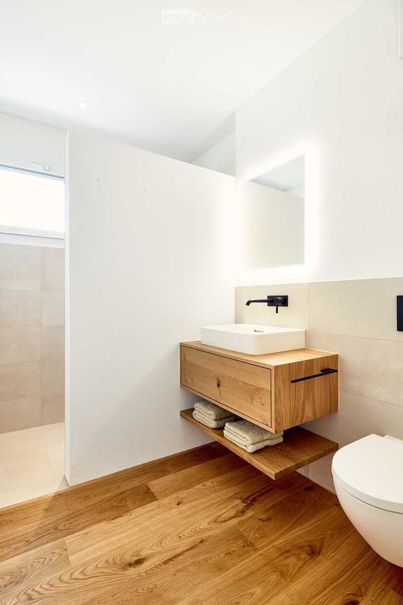 Modern bathroom with a wooden vanity, white sink, sleek faucet, and neatly folded towels on the shelf. The space features light wooden flooring and white walls.