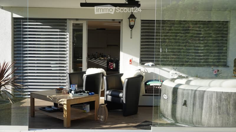 A modern outdoor patio with a wooden table, black leather sofa, hot tub, decorative vase, and potted plant. The space features large glass doors leading to an indoor area with a kitchen visible in the background.