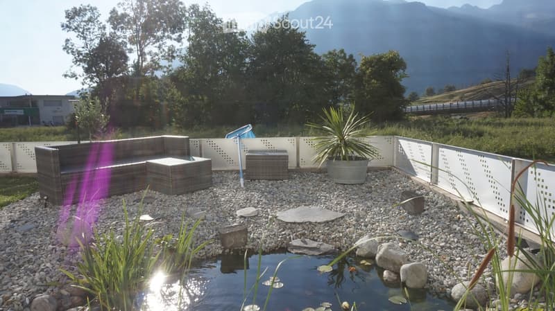 A modern outdoor seating area with a wicker sofa, table, and stools, surrounded by a small pond with rocks and plants, set against a scenic mountain backdrop.
