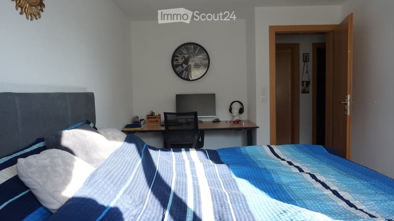 Bedroom with a blue and white striped bedspread, a grey headboard, a desk with a computer monitor, chair, books, and a toy car, along with a wall-mounted clock and a decorative sun-shaped wall piece.