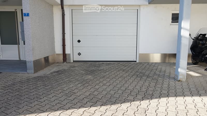 Modern building entrance with a large garage door, a paved driveway, and a scooter parked nearby.