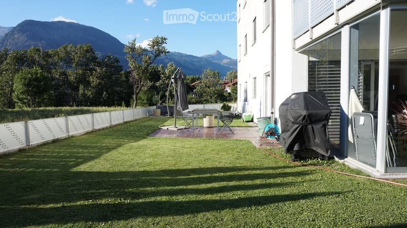 Well-maintained garden with artificial grass, patio area with table and chairs, covered grill, and scenic mountain view.