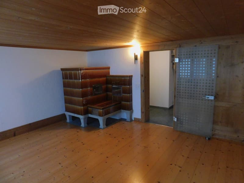 Empty room with wooden floor, white walls, and a built-in wooden bench with a tiled backrest. A doorway leads to another room, and a wall-mounted light fixture is visible.