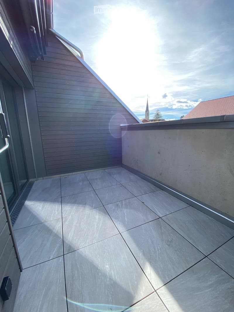 Modern balcony with tiled floor, wooden railing, and a view of a church spire in the distance.