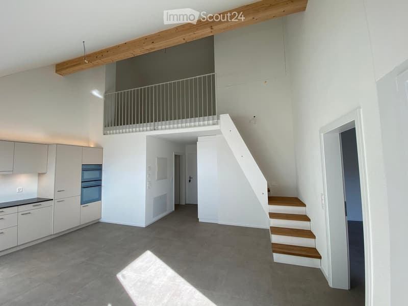 Open-plan living space with a modern kitchen area featuring white cabinets and an oven, a wooden staircase leading to an upper level, and a visible balcony railing above.